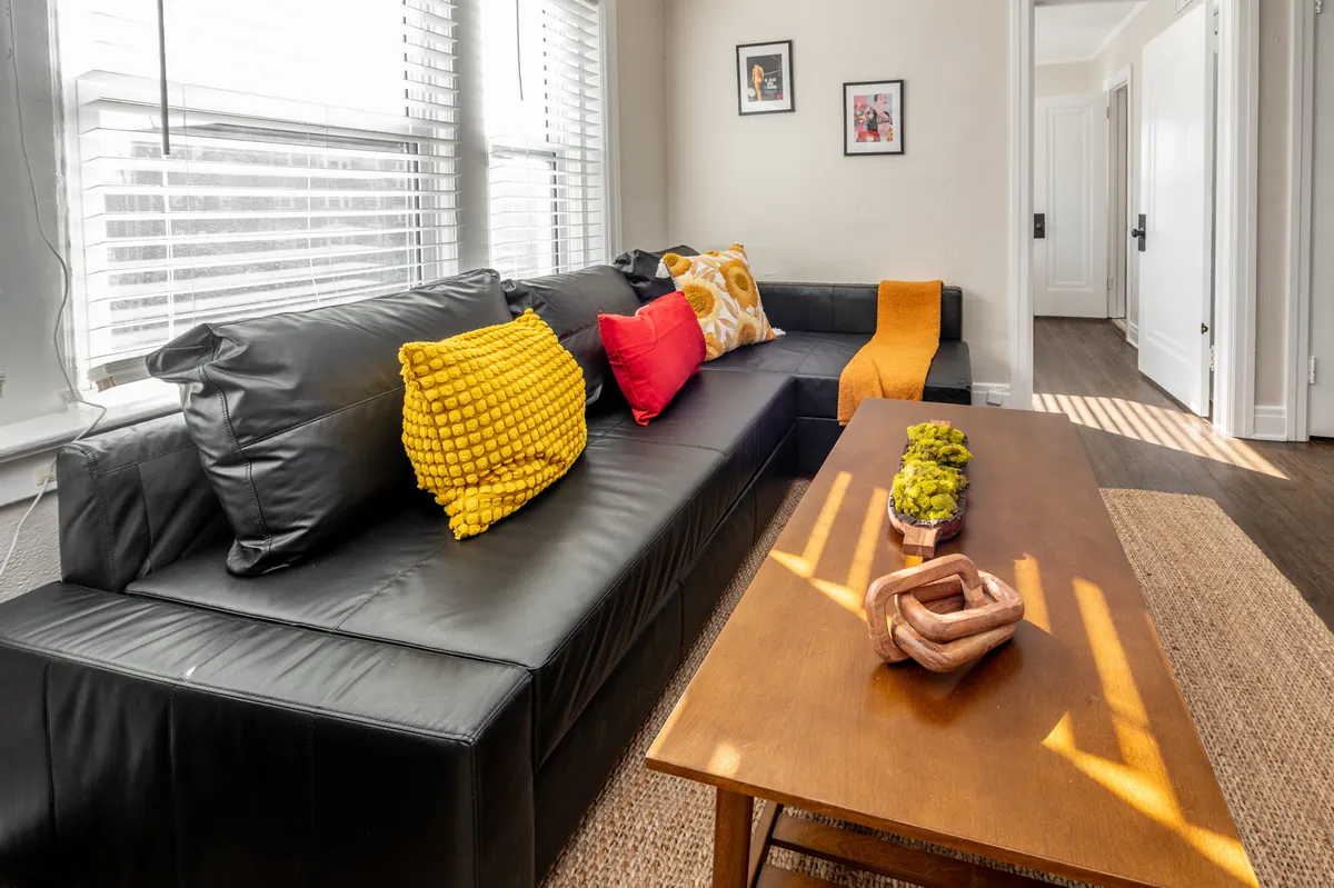 This image features a modern living space with a black leather sofa, colorful throw pillows, a wooden coffee table, and decorative touches. The scene exudes warmth and is perfect for relaxing, socializing, or planning outings in the city.