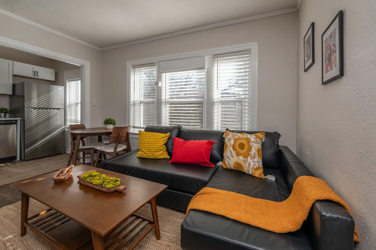 This image captures a living room scene featuring a black leather sofa adorned with colorful throw pillows, a wooden coffee table, and a view into a modern kitchen in the background. The space is infused with natural light and conveys a sense of comfort and relaxation, ideal for unwinding after a busy day.