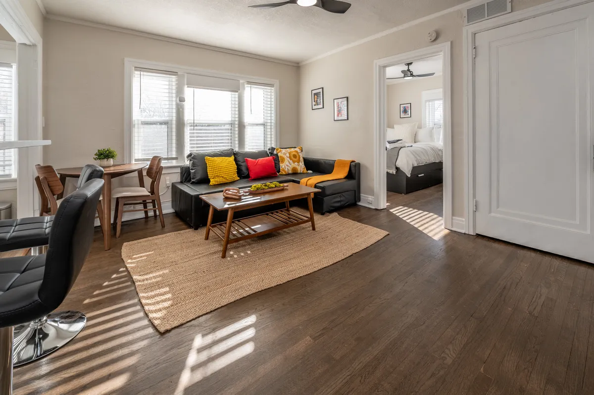 The image showcases a warm, inviting living space with a plush sofa adorned with colorful cushions, a stylish coffee table with decorative accents, and a small dining table perfect for intimate meals. The room feels bright and welcoming, suggesting comfort and togetherness in a cozy atmosphere.