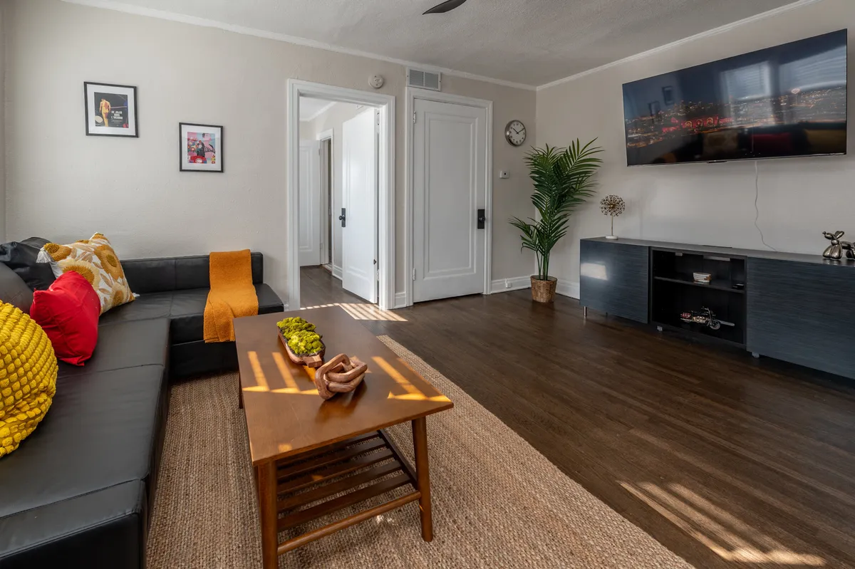 A modern living area with a black leather sofa, a stylish coffee table decorated with curated items, a potted plant, tasteful wall art, and a mounted TV. The overall mood is comfortable and contemporary, perfect for both socializing and relaxing solo.