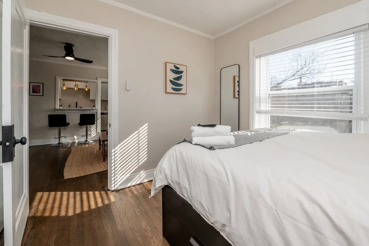 A calm and tidy bedroom scene with a neatly made bed, fresh towels, framed artwork, and abundant natural light streaming through a window. The ambiance is serene and inviting, suggesting a restful environment ideal for relaxation and quiet routines.